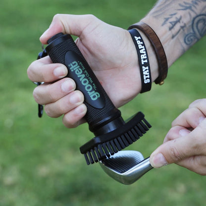 Person cleaning a golf club with a gooveit tool against a grassy background - Grooveit Brush – "The Wet Club Scrub" - Grooveit "The Wet Club Scrub" - #golf_club_cleaner# - #golf_club_brush# - #golf_brush# - #grooveit# - #groove_it_brush# - #grooveit_brush# - #groove_cleaner# - #golf_club_cleaning_brush# - #go